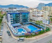 Relax Beach Hotel’in denize sıfır açık havuzu, palmiyelerle çevrili terası ve otel binasıyla birlikte güneşli bir yaz gününde görüntülenmiş