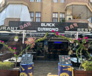 "Alanya’da deniz manzaralı Black Diamond Bar & Grill’in akşam saatlerinde aydınlatılmış açık hava oturma alanı ve ızgara servisi