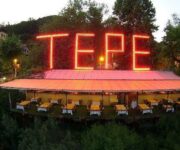 TEPE MOUNTANA RESTAURANT Alanya’da, yüksek rakımlı tepede kurulu terasta, Alanya manzarasına karşı akşam yemeği sofraları; masa üzerinde ızgara et, meze ve içecekler yer almakta.