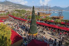 Noel Pazarı program detaylarını sergileyen bilgilendirme panosu ve etrafında toplanan ziyaretçilerin bulunduğu festival ortamı.”