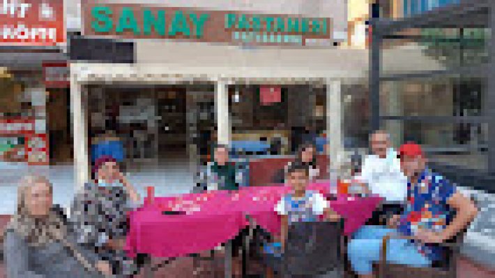 Sanay Pastanesi’nin vitrininde taze yaş pastalar, şerbetli tatlılar ve günlük çıkmış kurabiyeler sergileniyor, pastane içi ferah ve aydınlık bir şekilde görüntülenmiş