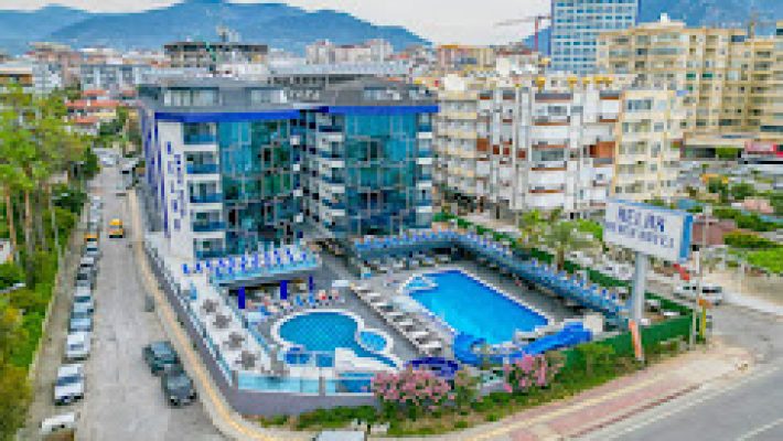 Relax Beach Hotel’in denize sıfır açık havuzu, palmiyelerle çevrili terası ve otel binasıyla birlikte güneşli bir yaz gününde görüntülenmiş