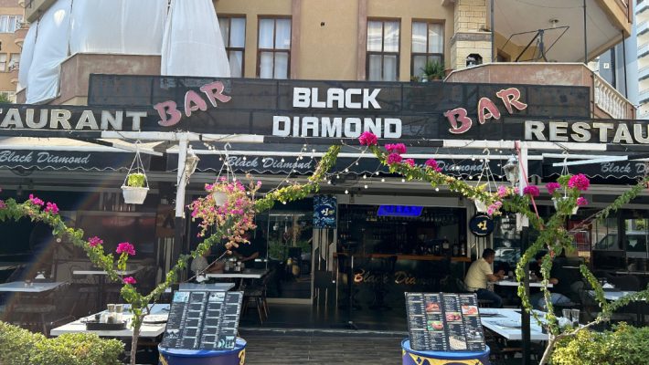 "Alanya’da deniz manzaralı Black Diamond Bar & Grill’in akşam saatlerinde aydınlatılmış açık hava oturma alanı ve ızgara servisi