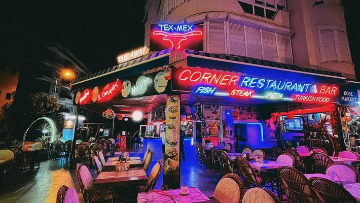 Corner Restaurant Alanya’da şık bir dış mekan masasında sunulmuş çeşitli deniz ürünleri, salatalar ve içecekler; arka planda Akdeniz manzarası ve yeşil bitkiler yer almakta.