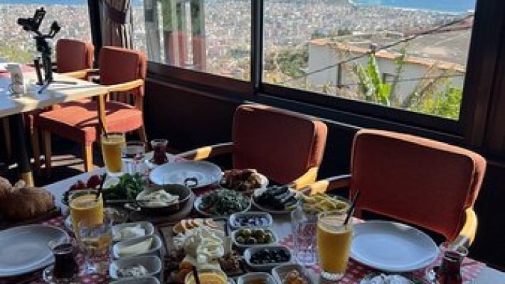 Kurşunlu Restaurant Alanya’da ağaçlarla çevrili, panoramik şehir ve deniz manzarasına sahip dış mekân; masalarda zengin mezeler, ızgara kebap tabakları ve içecekler sunulmaktadır.
