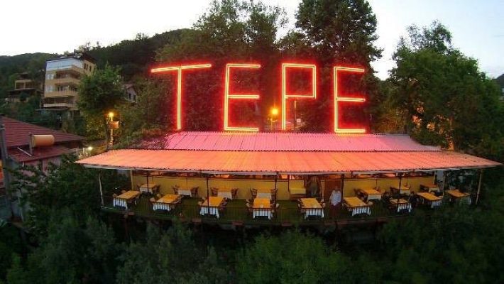TEPE MOUNTANA RESTAURANT Alanya’da, yüksek rakımlı tepede kurulu terasta, Alanya manzarasına karşı akşam yemeği sofraları; masa üzerinde ızgara et, meze ve içecekler yer almakta.